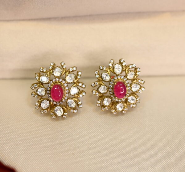 The image shows a pair of stud earrings. The earrings are gold-toned and have a floral design with a purple stone in the center. The design is intricate and detailed, with small white stones surrounding the center stone.