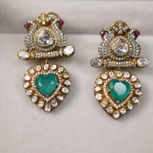 The image showcases a pair of elegant earrings. Shape: The earrings have a distinctive heart shape at the bottom, with a more intricate design at the top. Color: The heart-shaped pendants are a vibrant green, likely emeralds or green gemstones. They are surrounded by numerous smaller, colorless stones, probably diamonds or moissanites. Top Section: The top part of each earring features a more elaborate design with small, colorless stones arranged in a decorative pattern. There are also hints of pink stones visible.