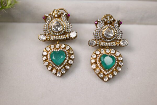 The image showcases a pair of elegant earrings. Shape: The earrings have a distinctive heart shape at the bottom, with a more intricate design at the top. Color: The heart-shaped pendants are a vibrant green, likely emeralds or green gemstones. They are surrounded by numerous smaller, colorless stones, probably diamonds or moissanites. Top Section: The top part of each earring features a more elaborate design with small, colorless stones arranged in a decorative pattern. There are also hints of pink stones visible.