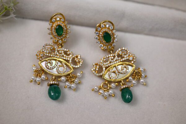 The image shows a pair of elegant earrings featuring a beautiful design with moissanite stones. The earrings appear to have a drop or teardrop shape, with a dangling element at the bottom. The main body of the earring seems to be crafted with a delicate filigree or latticework design. The design incorporates a central motif that resembles a stylized eye or a floral pattern.