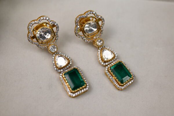 The image showcases a pair of elegant earrings. Each earring features a central rectangular emerald-cut moissanite, surrounded by a halo of smaller, sparkling white moissanites. Below the central stone, a pear-shaped moissanite dangles, adding a touch of movement and sophistication. The earrings appear to be crafted in gold, with intricate details surrounding the gemstones
