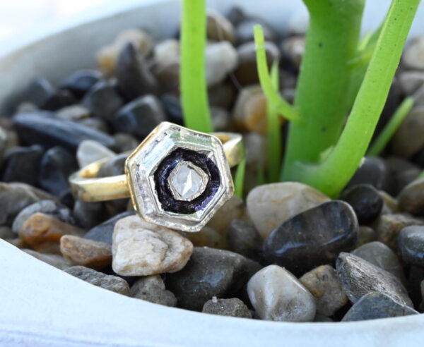 The image shows a close-up of a ring resting on a bed of small pebbles. The ring has a hexagonal setting with a prominent rose-cut diamond in the center. The diamond is surrounded by a delicate border of alternating black and white elements, creating a striking contrast. The ring itself appears to be made of gold or a similar metallic material. The background is blurred and features a small potted plant with green leaves.