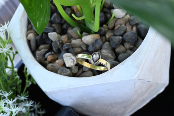 The image shows a close-up of a delicate ring resting on a bed of small, smooth stones. The ring features a single pear-shaped diamond set in a simple band. The band appears to be made of gold. The stones beneath the ring are a mix of light and dark colors, creating a natural and earthy backdrop. The background is blurred, drawing attention to the ring as the focal point of the image.