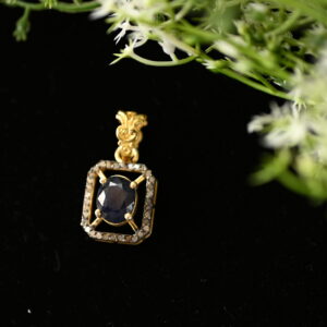 The image shows a close-up of a delicate gold pendant. The pendant features a large, oval-shaped blue sapphire as the centerpiece. The sapphire is surrounded by a halo of smaller, sparkling diamonds, creating a dazzling effect. The pendant itself has a simple, elegant design with intricate details on the bail (the part that connects to the chain). The background is a dark, velvety surface that contrasts beautifully with the golden color of the pendant.