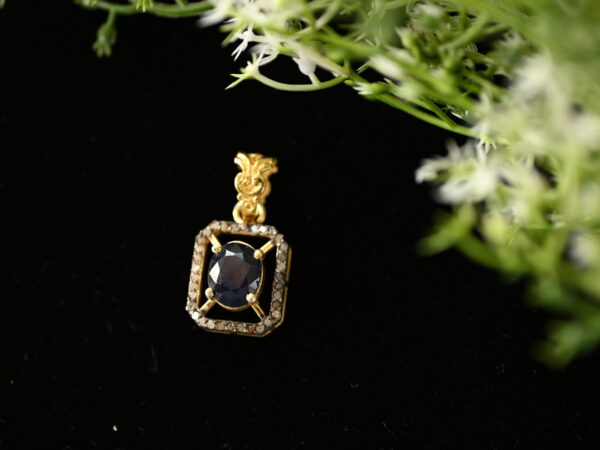 The image shows a close-up of a delicate gold pendant. The pendant features a large, oval-shaped blue sapphire as the centerpiece. The sapphire is surrounded by a halo of smaller, sparkling diamonds, creating a dazzling effect. The pendant itself has a simple, elegant design with intricate details on the bail (the part that connects to the chain). The background is a dark, velvety surface that contrasts beautifully with the golden color of the pendant.