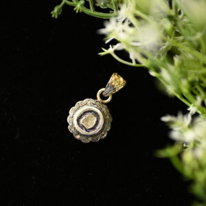 The image shows a close-up of a delicate gold pendant with a single diamond set in a scalloped bezel. The pendant is resting on a bed of dark fabric, with a sprig of green foliage and tiny white flowers partially obscuring the background.