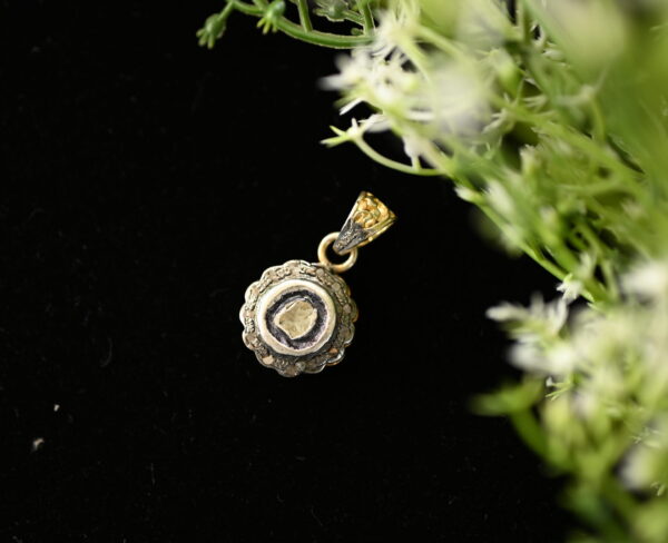 The image shows a close-up of a delicate gold pendant with a single diamond set in a scalloped bezel. The pendant is resting on a bed of dark fabric, with a sprig of green foliage and tiny white flowers partially obscuring the background.
