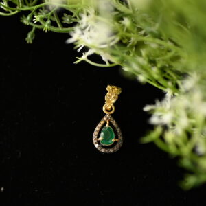 The image showcases a delicate and elegant gold pendant. The pendant features a pear-shaped emerald as its centerpiece, which is a vibrant shade of green. The emerald is surrounded by a halo of sparkling diamonds, creating a dazzling effect. The pendant is suspended from a gold chain, adding to its luxurious appeal. The background of the image is a dark surface, possibly velvet, which provides a striking contrast to the golden hues of the pendant.