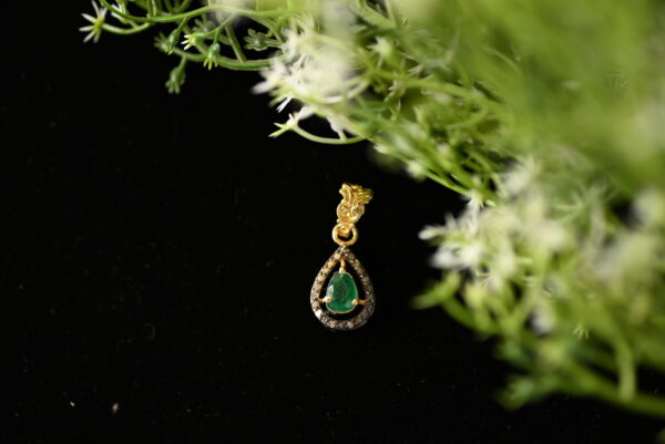 The image showcases a delicate and elegant gold pendant. The pendant features a pear-shaped emerald as its centerpiece, which is a vibrant shade of green. The emerald is surrounded by a halo of sparkling diamonds, creating a dazzling effect. The pendant is suspended from a gold chain, adding to its luxurious appeal. The background of the image is a dark surface, possibly velvet, which provides a striking contrast to the golden hues of the pendant.
