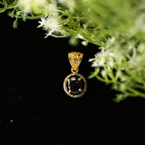 The image shows a gold pendant with a dark blue gemstone, likely a sapphire, set in a delicate gold frame adorned with tiny diamonds. The pendant is resting on a black surface, partially obscured by a sprig of white flowers and green leaves.
