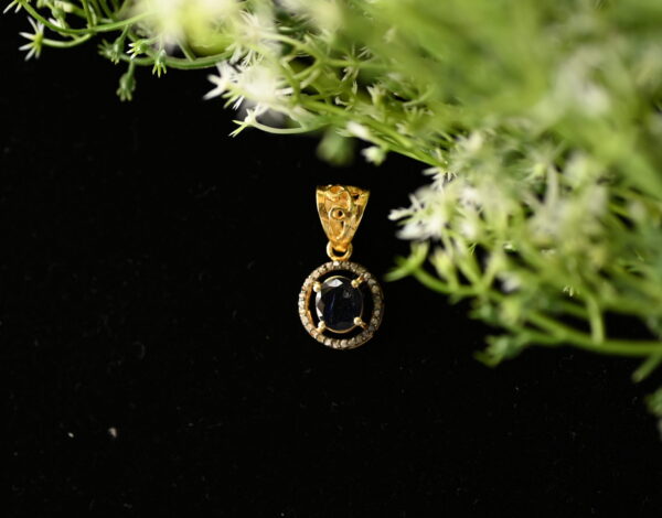 The image shows a gold pendant with a dark blue gemstone, likely a sapphire, set in a delicate gold frame adorned with tiny diamonds. The pendant is resting on a black surface, partially obscured by a sprig of white flowers and green leaves.