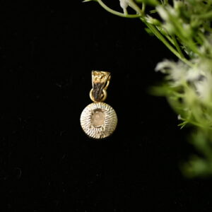 The image shows a close-up of a delicate gold pendant hanging against a dark background. A small sprig of greenery with white flowers is placed behind the pendant, adding a touch of nature to the scene. The pendant itself is circular with a textured edge and a central stone that appears to be a diamond.