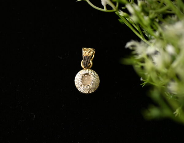 The image shows a close-up of a delicate gold pendant hanging against a dark background. A small sprig of greenery with white flowers is placed behind the pendant, adding a touch of nature to the scene. The pendant itself is circular with a textured edge and a central stone that appears to be a diamond.