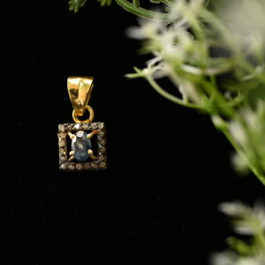 The image shows a close-up of a delicate gold pendant hanging against a dark background. The pendant features a square frame embellished with tiny diamonds, enclosing a single, round, dark blue gemstone. The background is a soft black, with a few slender green stems and white flowers partially visible in the top right corner. The flowers add a touch of nature to the image and create a contrast with the sleekness of the pendant.