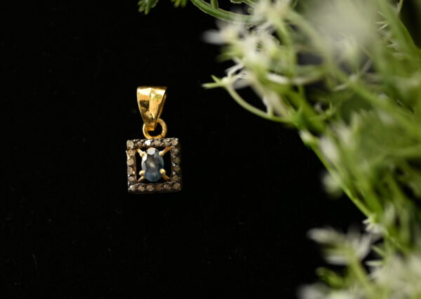 The image shows a close-up of a delicate gold pendant hanging against a dark background. The pendant features a square frame embellished with tiny diamonds, enclosing a single, round, dark blue gemstone. The background is a soft black, with a few slender green stems and white flowers partially visible in the top right corner. The flowers add a touch of nature to the image and create a contrast with the sleekness of the pendant.