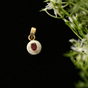 The image shows a close-up of a small, round pendant. Gemstone: A single, oval-shaped red gemstone is set in the center of the pendant. It has a smooth, polished surface. Background: The pendant is lying on a dark, possibly black, surface. Foliage: A few delicate green stems with tiny white flowers are partially visible in the background, adding a touch of nature to the scene.