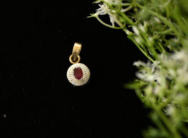 The image shows a close-up of a small, round pendant. Gemstone: A single, oval-shaped red gemstone is set in the center of the pendant. It has a smooth, polished surface. Background: The pendant is lying on a dark, possibly black, surface. Foliage: A few delicate green stems with tiny white flowers are partially visible in the background, adding a touch of nature to the scene.