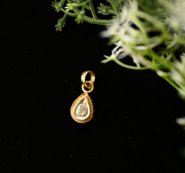 The image shows a close-up of a delicate gold pendant hanging from a thin chain. The pendant is shaped like a teardrop and holds a single, round diamond in its center. The diamond is clear and sparkling, reflecting light beautifully. The background of the image is dark, creating a dramatic contrast that highlights the pendant's elegance.