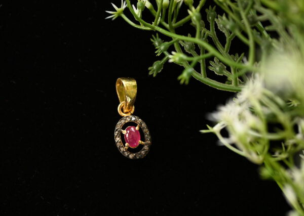 The image shows a close-up of a delicate pendant necklace. The pendant features a round, bezel-set ruby gemstone as the centerpiece. Surrounding the ruby is a halo of smaller, pave-set diamonds, creating a sparkling effect. The pendant itself appears to be crafted from 925 sterling silver. Background: The pendant is photographed against a dark background, possibly black velvet, which helps to highlight its details and make the gemstones shine.