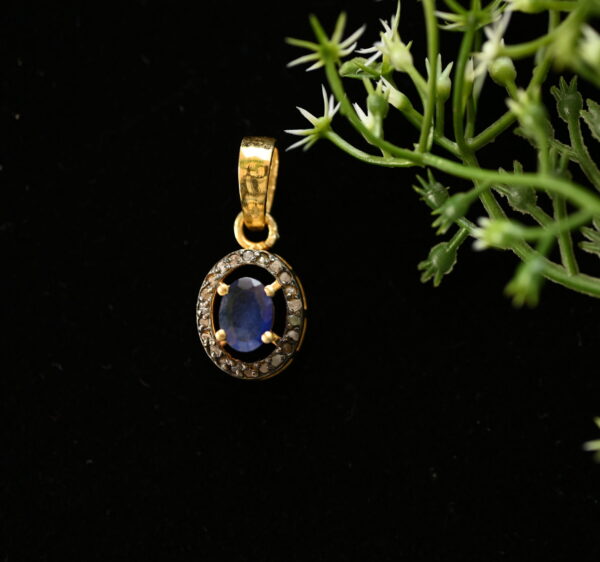 The image shows a beautiful pendant necklace. It features a round, deep blue sapphire gemstone set in a delicate gold frame. Surrounding the sapphire are several smaller diamonds, adding sparkle and brilliance to the piece. The pendant hangs from a simple gold chain. The background is a dark, possibly black, surface. There are some small green leaves and white flowers in the background, adding a touch of nature to the image.