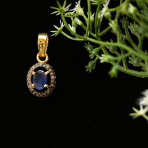 The image shows a delicate pendant necklace on a black background. The pendant is made of gold and features a central oval-shaped blue sapphire surrounded by a halo of smaller diamonds. The pendant hangs from a thin gold chain. The background is black, which makes the pendant stand out and draws attention to its intricate design.