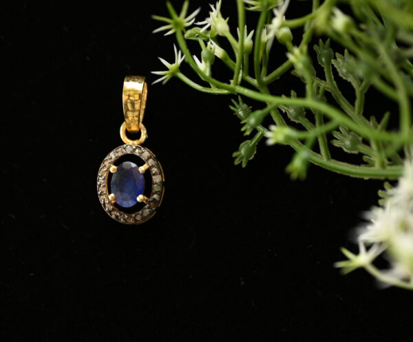 The image shows a delicate pendant necklace on a black background. The pendant is made of gold and features a central oval-shaped blue sapphire surrounded by a halo of smaller diamonds. The pendant hangs from a thin gold chain. The background is black, which makes the pendant stand out and draws attention to its intricate design.