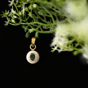 The image shows a gold pendant with a green gemstone set in the center. The pendant is hanging from a small branch with delicate green leaves and white flowers. The background is black.