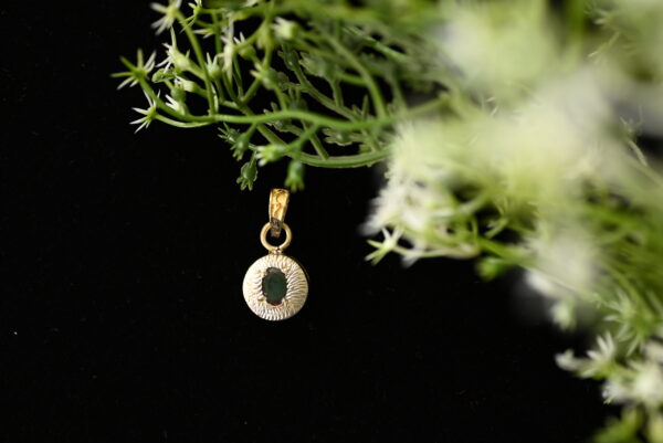 The image shows a gold pendant with a green gemstone set in the center. The pendant is hanging from a small branch with delicate green leaves and white flowers. The background is black.