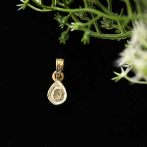 The image shows a close-up of a delicate diamond pendant. The pendant is teardrop-shaped and features a single, sparkling diamond set in a prong setting. It appears to be made of gold or yellow gold. The background is black, which helps to highlight the pendant's brilliance.
