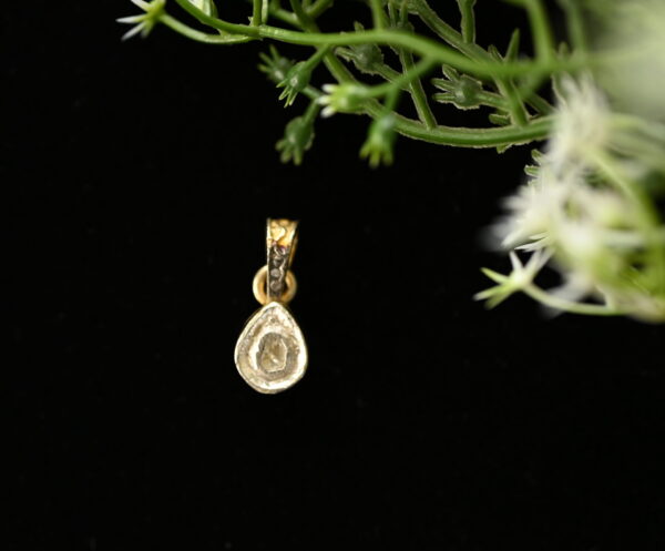 The image shows a close-up of a delicate diamond pendant. The pendant is teardrop-shaped and features a single, sparkling diamond set in a prong setting. It appears to be made of gold or yellow gold. The background is black, which helps to highlight the pendant's brilliance.