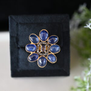 The image shows a close-up of a ring with a floral design. The ring features a cluster of oval-shaped blue sapphires arranged in a flower-like pattern. The sapphires are a deep, rich blue color.