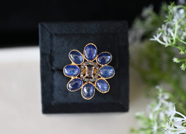 The image shows a close-up of a ring with a floral design. The ring features a cluster of oval-shaped blue sapphires arranged in a flower-like pattern. The sapphires are a deep, rich blue color.
