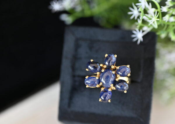 The image shows a close-up of a ring with a floral design. The ring is made of gold and features a cluster of blue sapphires set in a delicate pattern. The background is blurred and shows a few white flowers.