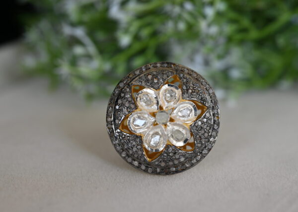 The image shows a close-up of a ring. The ring appears to be made of silver and has a circular design with a floral pattern. The center of the flower is filled with small diamonds, and the petals are outlined with smaller diamonds as well. The background of the image is blurred, with a green leafy texture visible.