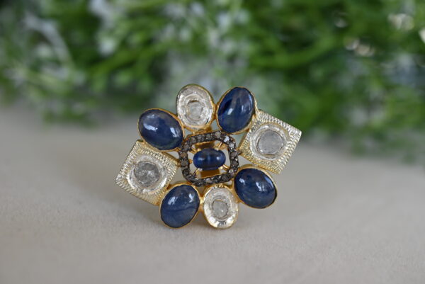The image shows a close-up of a ring with a unique design. It appears to be made of silver or a similar metal, with a central cluster of white diamonds. Surrounding the diamonds, there is a ring of deep blue sapphires. The overall design is geometric and intricate, with the diamonds and sapphires set in a patterned arrangement.