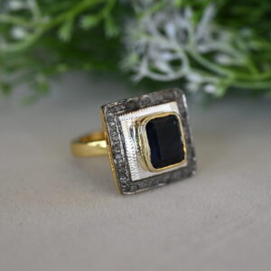 The image shows a close-up of a ring with a large, square-cut black gemstone set in a gold frame. The frame is embellished with smaller diamonds, creating a glittering border. The ring is resting on a surface with a blurred green background, possibly foliage or a plant.