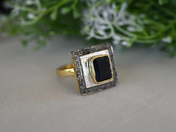 The image shows a close-up of a ring with a large, square-cut black gemstone set in a gold frame. The frame is embellished with smaller diamonds, creating a glittering border. The ring is resting on a surface with a blurred green background, possibly foliage or a plant.