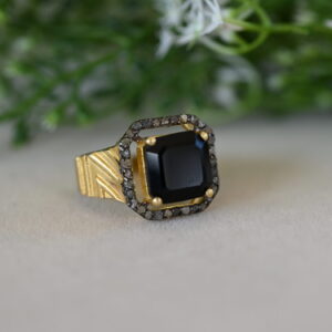 The image shows a close-up of a gold ring with a square-shaped black onyx gemstone in the center. The onyx is surrounded by a halo of small, sparkling diamonds. The ring band is textured with a gold-colored pattern. The ring is resting on a light-colored surface with a blurred background of green foliage and white flowers.