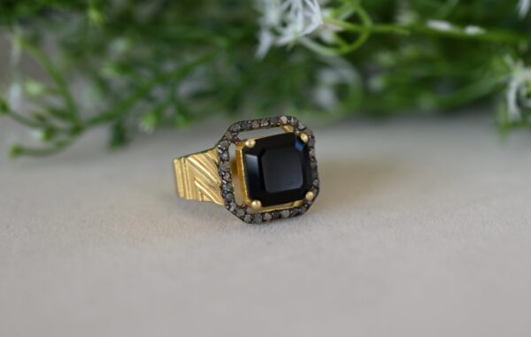 The image shows a close-up of a gold ring with a square-shaped black onyx gemstone in the center. The onyx is surrounded by a halo of small, sparkling diamonds. The ring band is textured with a gold-colored pattern. The ring is resting on a light-colored surface with a blurred background of green foliage and white flowers.