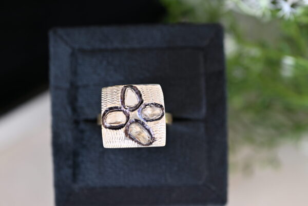 The image shows a close-up of a ring resting on a black velvet cushion inside a square box. The ring has a square-shaped setting with four raw diamonds set within it. The diamonds are irregular in shape and have a rough, unpolished appearance. The ring band is textured and appears to be made of gold. The background is blurred and shows a sprig of green foliage.