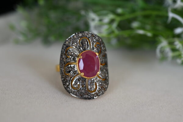 The image shows a close-up of a stunning ring. It appears to be made of gold and features a large, oval-shaped ruby as the centerpiece. The ruby is surrounded by a halo of smaller diamonds, creating a dazzling effect. The ring itself has intricate floral designs and a delicate filigree pattern, suggesting a vintage or antique style. The background of the image is blurred, with hints of green foliage visible, possibly suggesting the ring is resting on a bed of leaves or flowers.