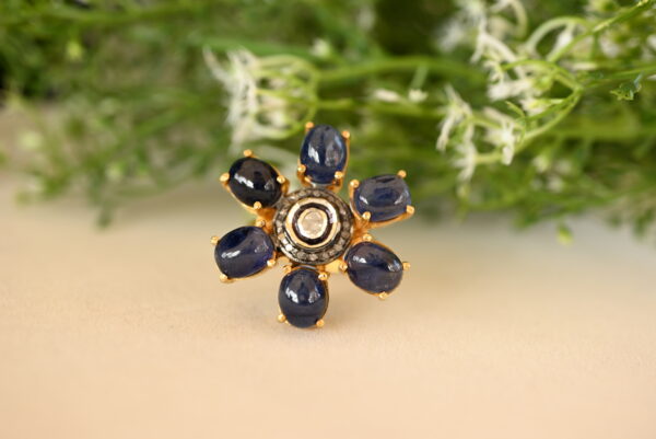 The image shows a beautiful ring with a floral design. It appears to be made of gold and features a cluster of seven deep blue sapphires arranged in a flower-like pattern. In the center of the flower, there is a single, sparkling diamond. The ring is resting on a surface that looks like a piece of fabric, with some green foliage in the background.
