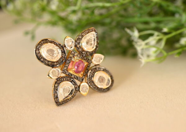 The image shows a close-up of a beautiful diamond ring. The ring appears to be made of 925 silver and is gold-plated. It features a central pink gemstone, likely a ruby, surrounded by a cluster of sparkling diamonds. The design is intricate, with a floral motif that adds to its elegance. The ring is resting on a surface with a blurred background of green foliage, possibly a plant or a bouquet.
