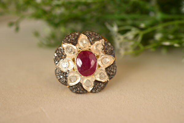 The image shows a close-up of a beautiful ring resting on a textured surface. The ring is adorned with a large, oval-shaped ruby in the center, surrounded by a cluster of smaller diamonds. The background of the image is blurred, with hints of green foliage visible, suggesting a natural setting. Radiant