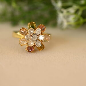 The image shows a close-up of a gold ring with a floral design. The ring is set with a cluster of small white diamonds in the center, surrounded by a ring of larger, colorful gemstones. The gemstones appear to be pink sapphires, yellow citrines, and green emeralds. The ring is resting on a beige surface with a blurred background of green foliage.