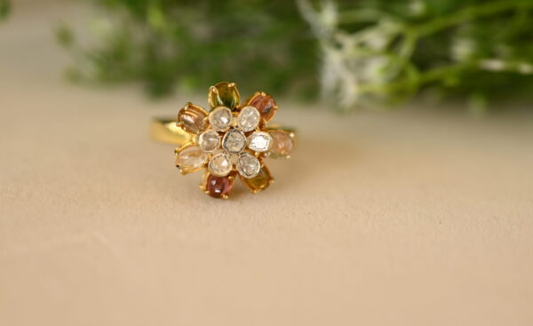 The image shows a close-up of a gold ring with a floral design. The ring is set with a cluster of small white diamonds in the center, surrounded by a ring of larger, colorful gemstones. The gemstones appear to be pink sapphires, yellow citrines, and green emeralds. The ring is resting on a beige surface with a blurred background of green foliage.