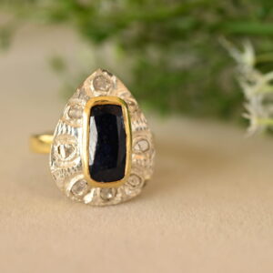 The image shows a close-up of a ring resting on a surface with a blurred background of green foliage. The ring appears to be made of silver or white gold with a teardrop-shaped setting. Inside the setting is a rectangular dark blue gemstone, likely a sapphire. The teardrop setting is embellished with smaller diamonds, creating a halo effect. The band of the ring has intricate, textured detailing.