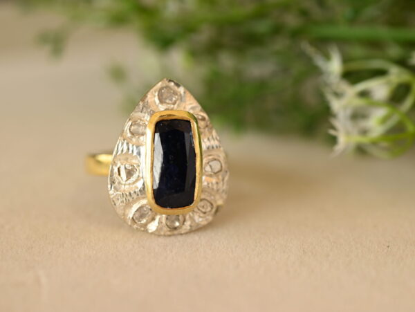 The image shows a close-up of a ring resting on a surface with a blurred background of green foliage. The ring appears to be made of silver or white gold with a teardrop-shaped setting. Inside the setting is a rectangular dark blue gemstone, likely a sapphire. The teardrop setting is embellished with smaller diamonds, creating a halo effect. The band of the ring has intricate, textured detailing.