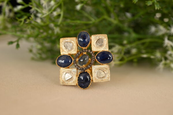 The image shows a close-up of a ring. The ring appears to be made of silver and features a central square design. Within the square, there are several round blue sapphires and smaller diamonds. The diamonds are set in a way that creates a sparkling halo around the sapphires.