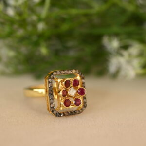 The image shows a close-up of a gold-toned ring with a square-shaped center. The center is filled with small red rubies and a single white diamond. The ring is surrounded by a delicate diamond border, and the background is blurred with green foliage.